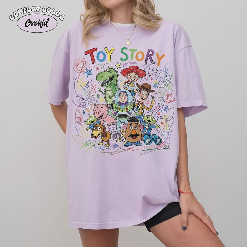 May include: A light purple Comfort Colors t-shirt featuring the words "Toy Story" in colorful letters. The shirt showcases a vibrant graphic of characters from the animated film, including Woody, Buzz Lightyear, and Rex.