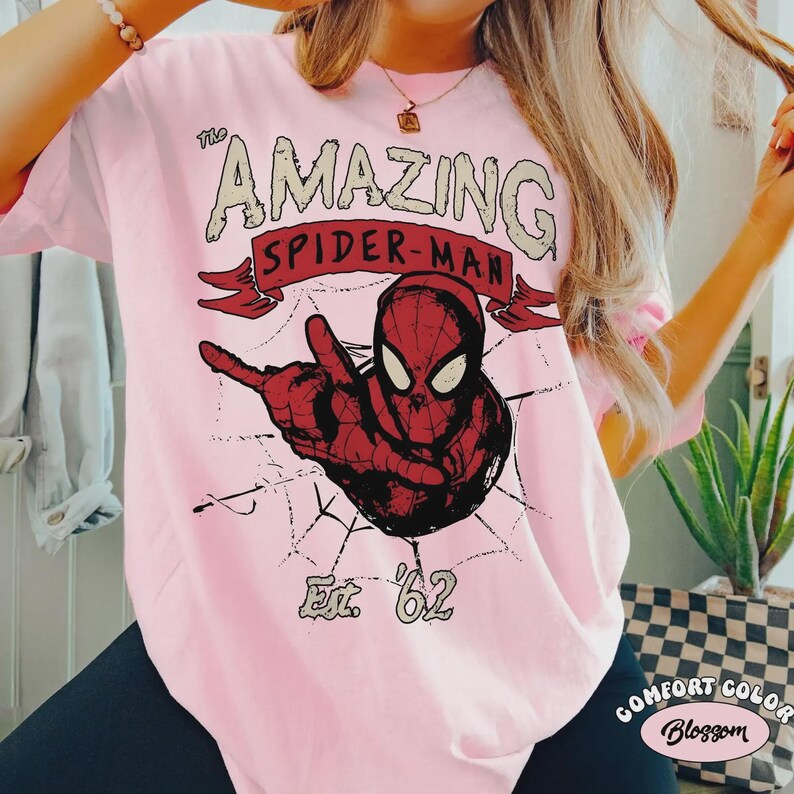 May include: Pink t-shirt featuring a red and black graphic of Spider-Man with the text "The Amazing Spider-Man" and "Est. '62". The design includes a spider web and is printed on a Comfort Colors shirt.