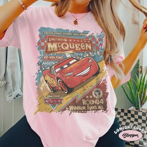 May include: Pink t-shirt featuring a graphic of Lightning McQueen from the Disney Pixar Cars movie. The design includes text that reads "Lightning McQueen", "Cars", and "Winner Takes All". The shirt is a Comfort Color brand.