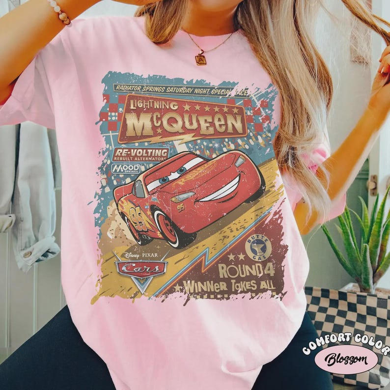May include: Pink t-shirt featuring a graphic of Lightning McQueen from the Disney Pixar Cars movie. The design includes text that reads "Lightning McQueen", "Cars", and "Winner Takes All". The shirt is a Comfort Color brand.