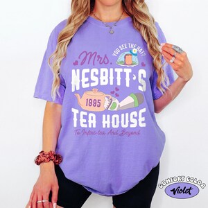 May include: A violet-colored t-shirt with the text "Mrs. Nesbitt's Tea House" and a teapot graphic. The shirt also features the text "You see the hat?" and "To Infini-tea And Beyond."
