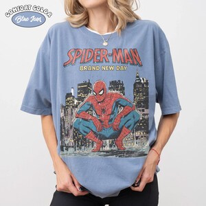 May include: A blue Comfort Colors t-shirt featuring a Spider-Man graphic. The design includes the words "SPIDER-MAN BRAND NEW DAY" above an illustration of Spider-Man crouching in front of a city skyline. The shirt has short sleeves.