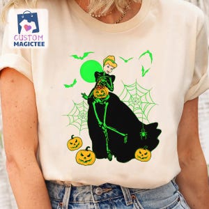 May include: Cream-colored t-shirt featuring a Halloween-themed graphic. The design includes a skeletonized princess holding a jack-o'-lantern, surrounded by green bats, spiderwebs, and pumpkins. The text "CUSTOM MAGICTEE" is in the upper left corner.