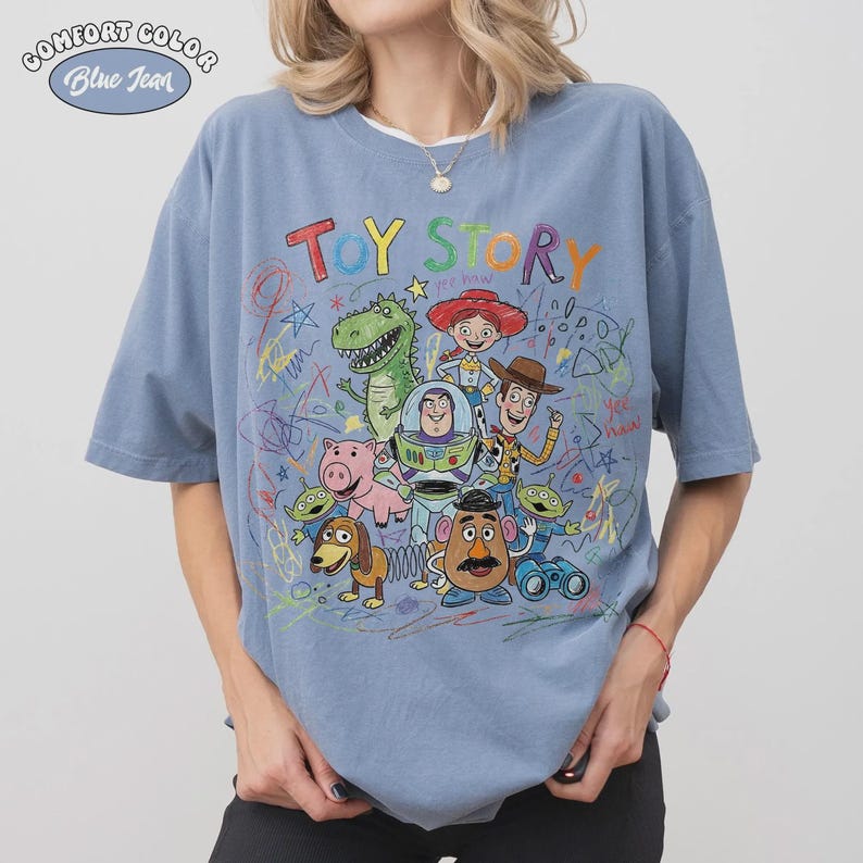 May include: Blue jean Comfort Colors t-shirt featuring a colorful graphic of Toy Story characters. The design includes Woody, Buzz Lightyear, and other characters from the animated film, with the words "Toy Story" in a playful font.