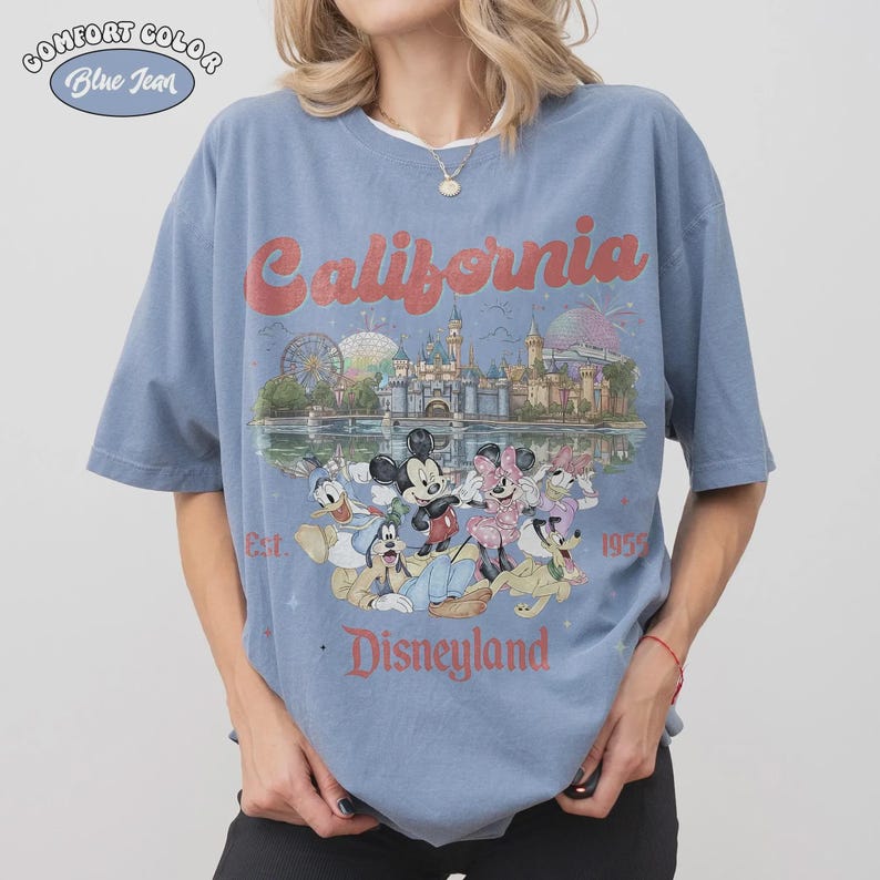 May include: Blue jean Comfort Colors t-shirt featuring a Disneyland California design. The design includes the text "California" in red script, a Disneyland castle, and cartoon characters. The shirt is a light blue color.