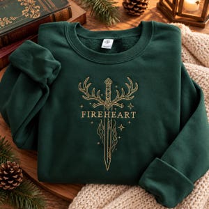 Peut inclure: Un sweat-shirt à col rond vert forêt foncé avec un motif brodé doré sur la poitrine. Le graphisme représente une épée centrale flanquée de bois de cerf, entourée de petites étoiles et de flammes, avec le texte FIREHEART centré en dessous.