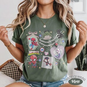 May include: Olive green t-shirt featuring cartoon illustrations of Spider-Man and friends. The design includes the text "Spidey and Amazing Friends" and other characters. The shirt is labeled "Comfort Color Moss".