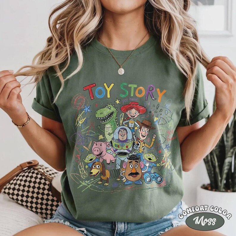 May include: Moss green t-shirt featuring the words "Toy Story" in colorful letters above a graphic of the movie's characters. The shirt has a crew neck and short sleeves. The words "Comfort Color" and "Moss" are printed on the bottom right.