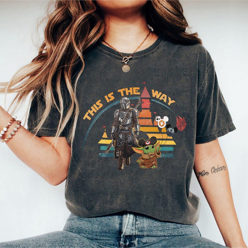 May include: A dark gray t-shirt with a vintage-style graphic of the Mandalorian and Grogu standing in front of a stylized version of the Cinderella Castle. The text "This is the way" is written in a circular arc around the graphic.