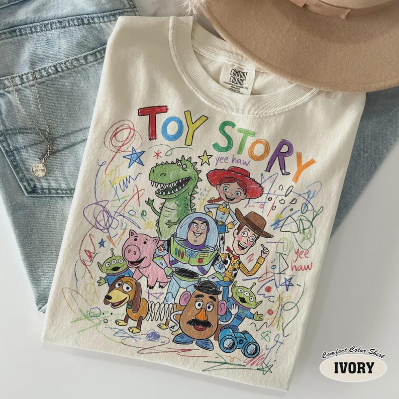 May include: Ivory-colored Comfort Colors t-shirt featuring a colorful, hand-drawn style graphic of characters from the movie "Toy Story." The design includes the words "Toy Story" in a playful font, along with various characters.