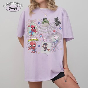 May include: A light purple t-shirt featuring cartoon images of Spider-Man and other characters from the Spidey and His Amazing Friends series. The shirt has the words "Spidey and Amazing Friends" printed on it.