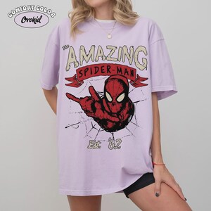 May include: A light purple t-shirt with the words "The Amazing Spider-Man" in a vintage style. The graphic features Spider-Man in red and black, with a spider web design and the text "Est. '62". The shirt has the text "Comfort Color Orchid" in the upper left corner.