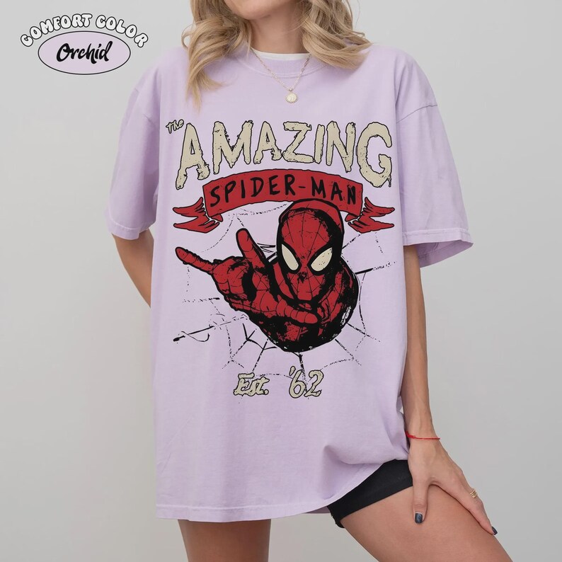 May include: A light purple t-shirt with the words "The Amazing Spider-Man" in a vintage style. The graphic features Spider-Man in red and black, with a spider web design and the text "Est. '62". The shirt has the text "Comfort Color Orchid" in the upper left corner.