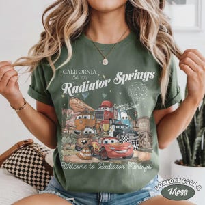 May include: Green t-shirt with a graphic of characters from the movie Cars, including Lightning McQueen, Mater, and others, with the text "California, Est. 2012, Radiator Springs, Welcome to Radiator Springs". The shirt is Comfort Color Moss.