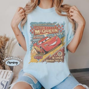 May include: Light blue t-shirt featuring Lightning McQueen from the Disney Pixar Cars movie. The graphic includes the text "Lightning McQueen", "Round 4", and "Winner Takes All". The shirt is labeled "Comfort Color Chambray".