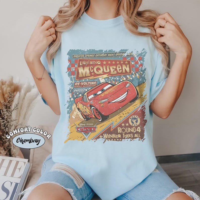 May include: Light blue t-shirt featuring Lightning McQueen from the Disney Pixar Cars movie. The graphic includes the text "Lightning McQueen", "Round 4", and "Winner Takes All". The shirt is labeled "Comfort Color Chambray".