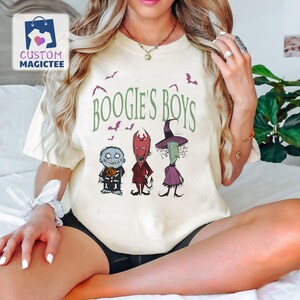 May include: Cream-colored t-shirt featuring the text "BOOGIE'S BOYS" in green, with illustrations of three characters. The characters include a blue-skinned figure, a red devil-like figure, and a green witch. Bats are also illustrated.