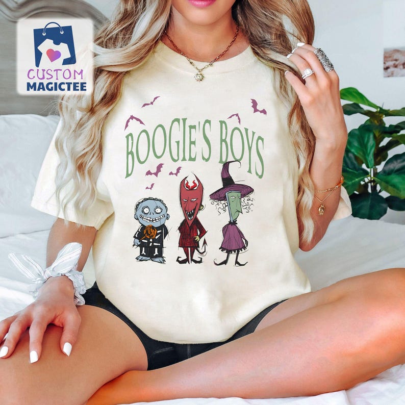 May include: Cream-colored t-shirt featuring the text "BOOGIE'S BOYS" in green, with illustrations of three characters. The characters include a blue-skinned figure, a red devil-like figure, and a green witch. Bats are also illustrated.