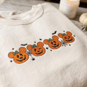 Puede incluir: Una sudadera de cuello redondo color crema con un diseño bordado de tres calabazas naranjas con orejas de ratón y caras de linterna. El bordado incluye pequeñas telarañas negras, arañas, murciélagos y estrellas.