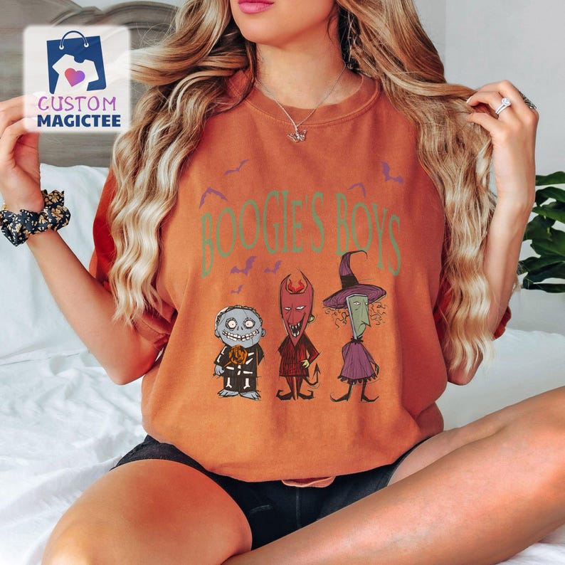 May include: Orange t-shirt featuring the text "BOOGIE'S BOYS" in green, with three cartoon characters below. The characters include a pumpkin-headed figure, a devil, and a witch. The shirt has a relaxed fit.