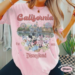 May include: Pink t-shirt with a graphic featuring Disneyland characters and the text "California Disneyland Est. 1955." The design includes Mickey and Minnie Mouse, Donald Duck, and Goofy, with a castle and Ferris wheel.