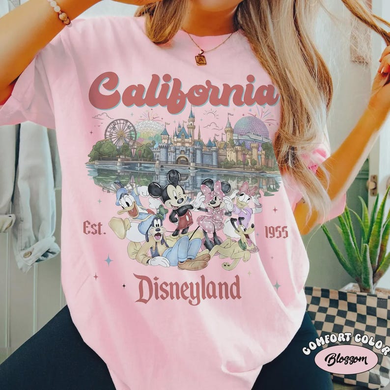 May include: Pink t-shirt with a graphic featuring Disneyland characters and the text "California Disneyland Est. 1955." The design includes Mickey and Minnie Mouse, Donald Duck, and Goofy, with a castle and Ferris wheel.