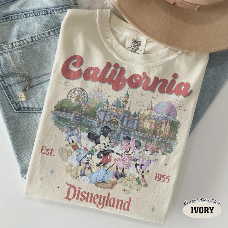 May include: Ivory-colored t-shirt with the word "California" in red script. The shirt features a Disneyland graphic with Mickey and Minnie Mouse, Donald Duck, and Goofy. The text "Est. 1955 Disneyland" is also printed on the shirt.