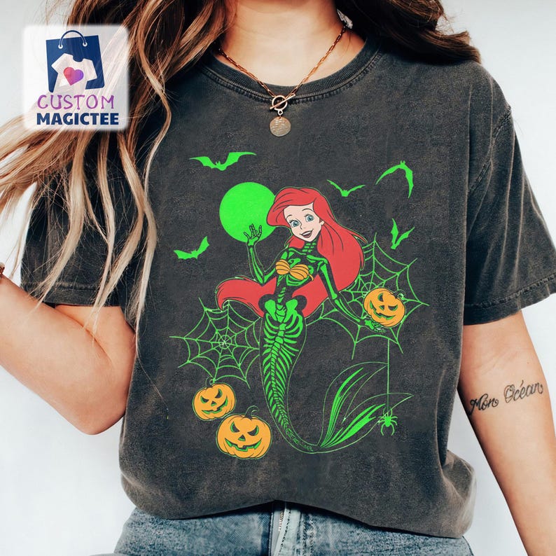 May include: Dark gray t-shirt featuring a Halloween-themed design. The graphic depicts a mermaid with a skeleton torso, holding a pumpkin, with a green moon and bats. The shirt also has spiderwebs and jack-o'-lanterns.