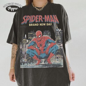 May include: Dark gray t-shirt featuring a graphic of Spider-Man in a crouching pose, with the words "SPIDER-MAN BRAND NEW DAY" in red and blue. The shirt has a vintage, distressed look, with the Comfort Colors Pepper logo in the upper left corner.