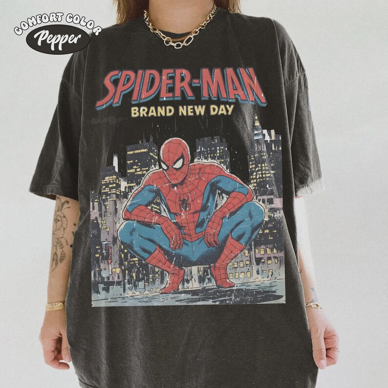 May include: Dark gray t-shirt featuring a graphic of Spider-Man in a crouching pose, with the words "SPIDER-MAN BRAND NEW DAY" in red and blue. The shirt has a vintage, distressed look, with the Comfort Colors Pepper logo in the upper left corner.