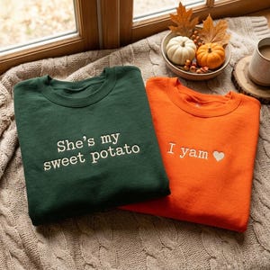 Könnte beinhalten: Ein dunkelgrünes Sweatshirt mit dem weißen gestickten Text 'She's my sweet potato' und ein oranges Sweatshirt mit dem weißen gestickten Text 'I yam' und einem kleinen weißen Herzsymbol, beide mit Rundhalsausschnitt und langen Ärmeln.