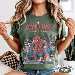May include: Green t-shirt featuring a vintage Spider-Man graphic. The design includes the words "Spider-Man Brand New Day" above an illustration of Spider-Man crouching in front of a cityscape. The shirt is labeled "Comfort Color Moss".