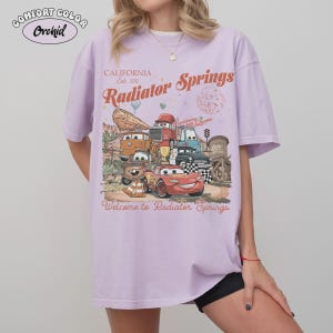 May include: A light purple Comfort Colors t-shirt featuring a graphic of characters from the movie Cars. The graphic includes the text "California Est. 2012 Radiator Springs" and "Welcome to Radiator Springs".