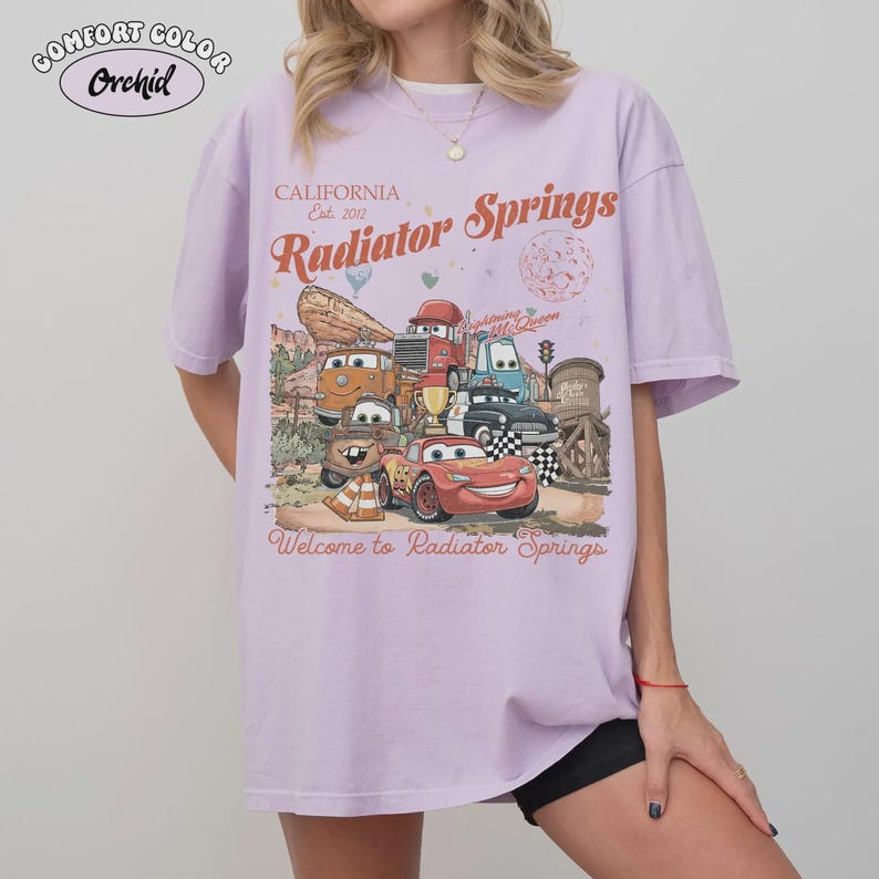 May include: A light purple Comfort Colors t-shirt featuring a graphic of characters from the movie Cars. The graphic includes the text "California Est. 2012 Radiator Springs" and "Welcome to Radiator Springs".
