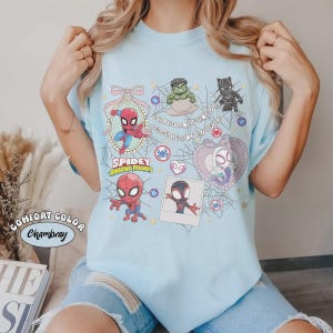 May include: Light blue Comfort Color Chambray t-shirt featuring Spidey and his Amazing Friends characters. The design includes Spider-Man, Hulk, Black Panther, and Spider-Gwen, with the text "Spidey Amazing Friends".