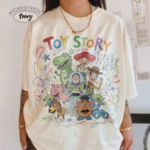 May include: Ivory Comfort Color t-shirt featuring a colorful Toy Story graphic. The design includes characters like Woody, Buzz Lightyear, and Rex, with the words "Toy Story" in a playful font. The shirt has a crew neck and short sleeves.
