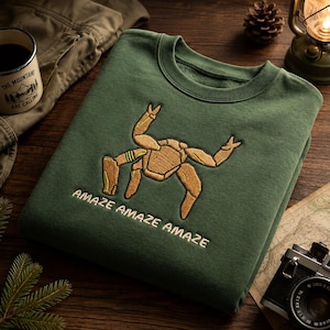 May include: A forest green crewneck sweatshirt featuring an embroidered tan crab-like creature with raised claws. Below the creature, the text "AMAZE AMAZE AMAZE" is embroidered in a light cream colour. The sweatshirt has a ribbed collar and cuffs.
