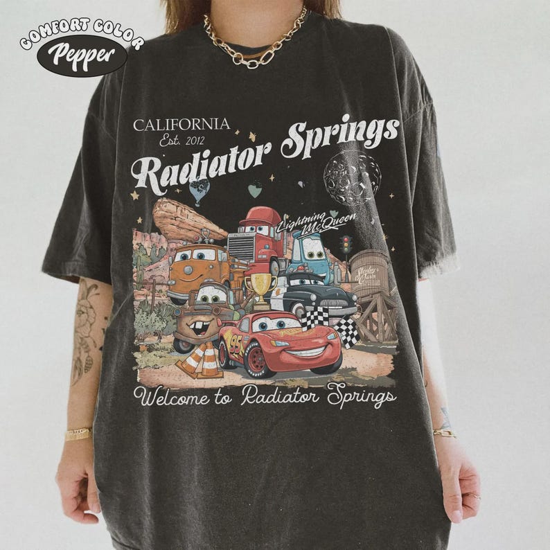May include: Dark gray t-shirt with a graphic featuring characters from the movie Cars, including Lightning McQueen. The shirt has the text "CALIFORNIA Est. 2012 Radiator Springs" and "Welcome to Radiator Springs". The shirt is labeled "Comfort Color Pepper".