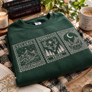 May include: A dark green crewneck sweatshirt featuring three white embroidered rectangular stamp-style designs. The designs depict mountain peaks labelled Velaris City of Starlight, a deer head with trees labelled Terrasen Erilea, and a crescent moon over a castle labelled Crescent City.