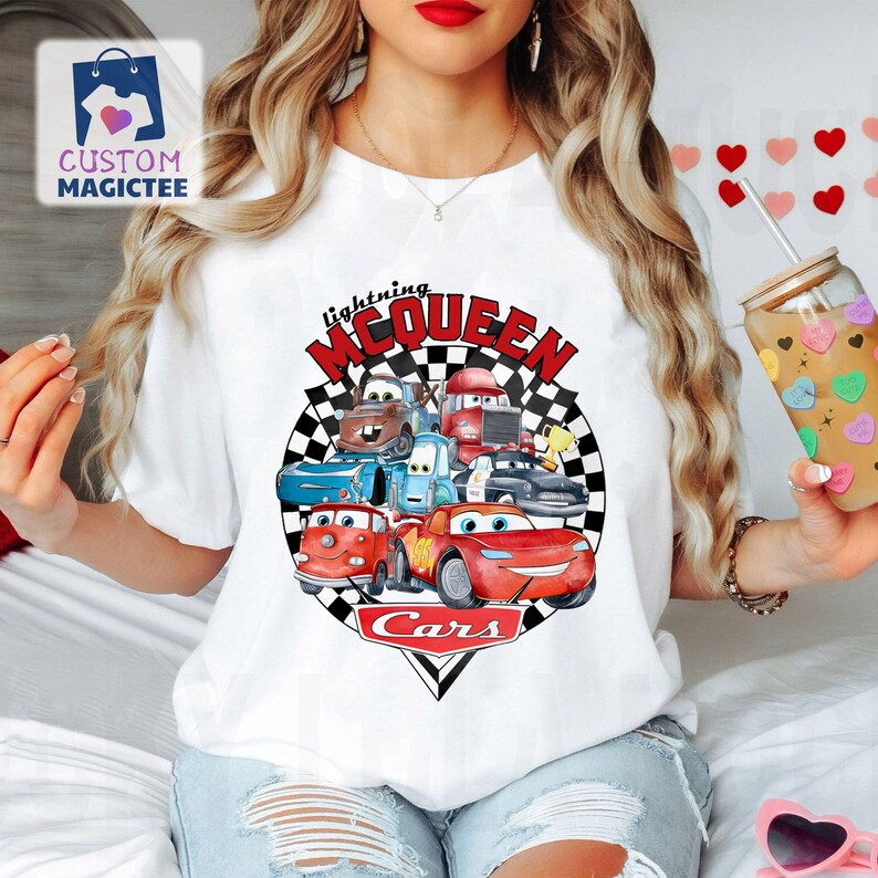 May include: White t-shirt with a colourful graphic of characters from the Cars film, including Lightning McQueen. The design features the text "Lightning McQueen" and "Cars" in red and black, set against a chequered background.