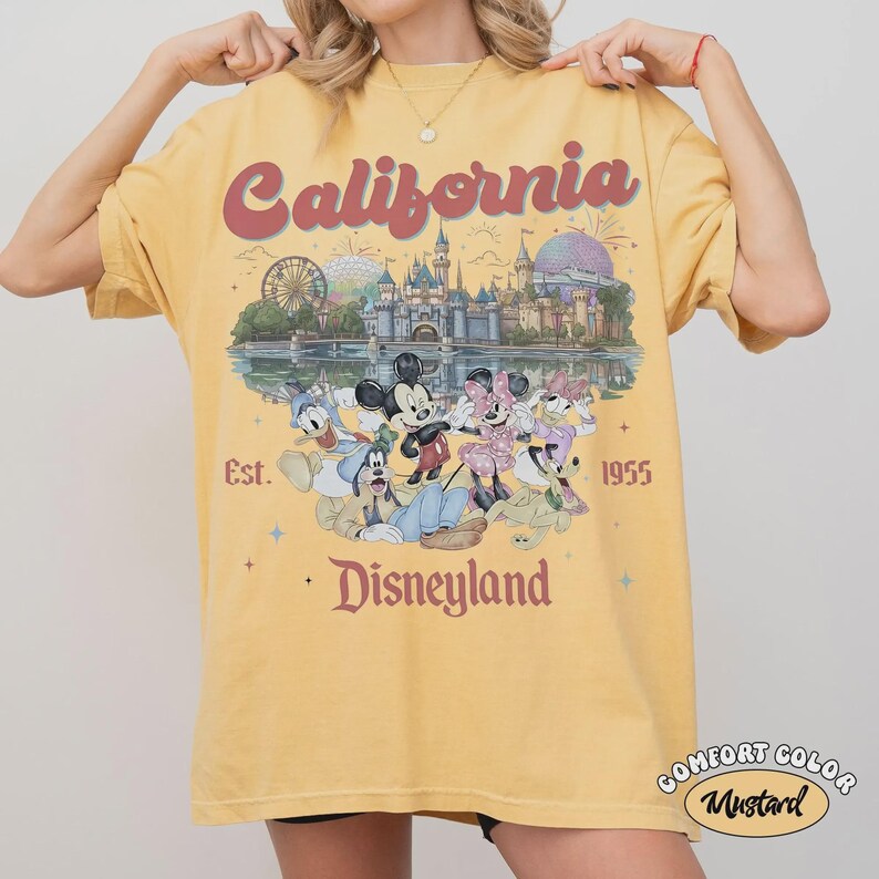 May include: Mustard yellow t-shirt with a graphic featuring the word "California" in red script above a Disneyland scene with Mickey and Minnie Mouse, Donald Duck, and Goofy. The text "Est. 1955" and "Disneyland" are also visible.