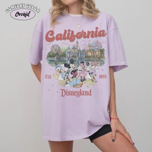 May include: A lilac Comfort Colors t-shirt featuring a Disneyland graphic. The design includes the word "California" in a retro font, the Disneyland castle, and various Disney characters. The text "Est. 1955" and "Disneyland" are also visible.