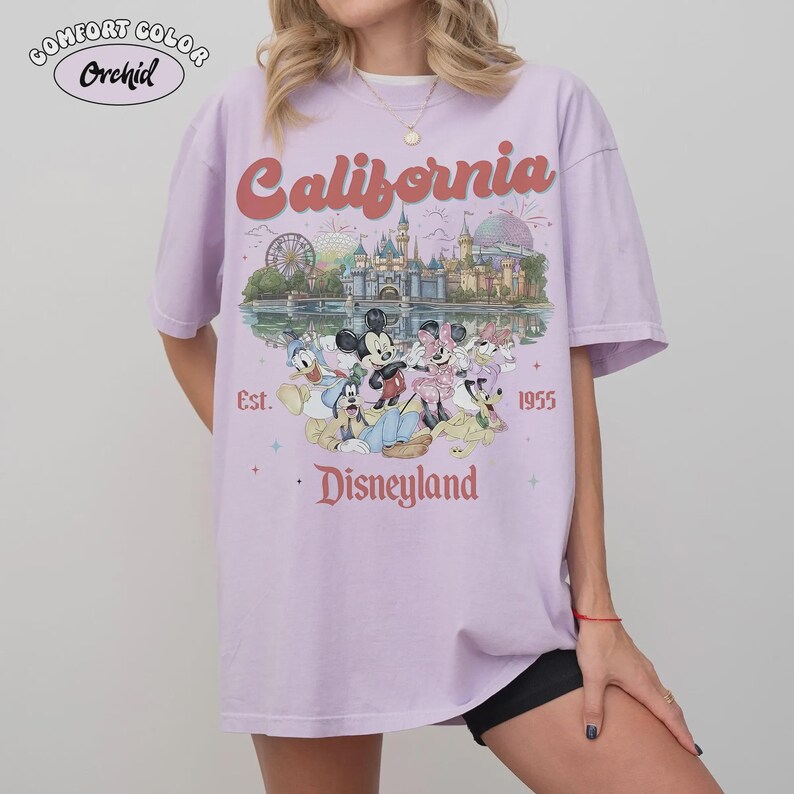 May include: A lilac Comfort Colors t-shirt featuring a Disneyland graphic. The design includes the word "California" in a retro font, the Disneyland castle, and various Disney characters. The text "Est. 1955" and "Disneyland" are also visible.