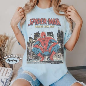 May include: Light blue t-shirt featuring a vintage-style Spider-Man graphic. The design includes the words "SPIDER-MAN BRAND NEW DAY" above an illustration of Spider-Man crouching in front of a city skyline. The shirt is labeled "Comfort Color Chambray".