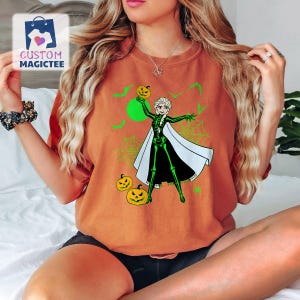 May include: Orange t-shirt featuring a Halloween-themed graphic. The design includes a cartoon character in a skeleton costume holding a jack-o'-lantern, with green accents, bats, and spiderwebs. The shirt has the text "CUSTOM MAGICTEE" in the upper left corner.