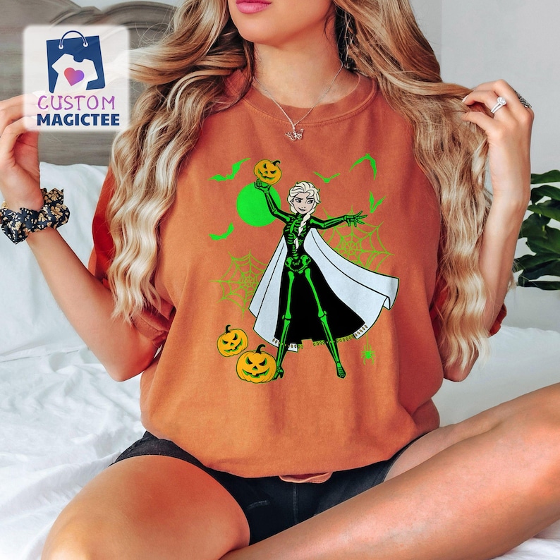 May include: Orange t-shirt featuring a Halloween-themed graphic. The design includes a cartoon character in a skeleton costume holding a jack-o'-lantern, with green accents, bats, and spiderwebs. The shirt has the text "CUSTOM MAGICTEE" in the upper left corner.