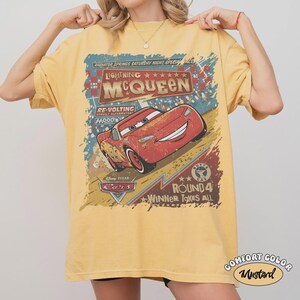 May include: Mustard yellow t-shirt featuring Lightning McQueen from the Disney Pixar Cars movie. The graphic includes text that reads "Lightning McQueen" and "Round 4 Winner Takes All". The shirt is a Comfort Color brand.