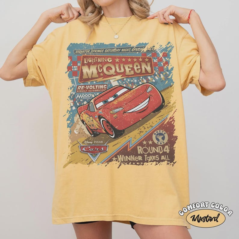 May include: Mustard yellow t-shirt featuring Lightning McQueen from the Disney Pixar Cars movie. The graphic includes text that reads "Lightning McQueen" and "Round 4 Winner Takes All". The shirt is a Comfort Color brand.