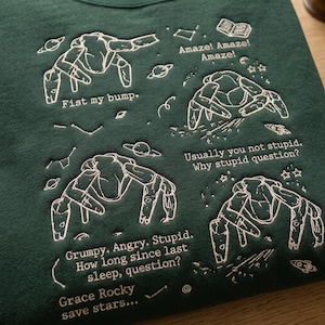 May include: A dark green sweatshirt featuring white embroidered line art of four alien creatures surrounded by space-themed icons like planets, stars, and rockets. The text includes phrases such as Fist my bump, Amaze! Amaze! Amaze!, Usually you not stupid. Why stupid question?, Grumpy. Angry. Stupid. How long since last sleep, question?, and Grace Rocky save stars...
