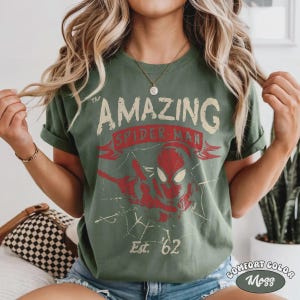May include: Green t-shirt with a distressed graphic of Spider-Man. The design includes the words "The Amazing Spider-Man" and "Est. '62" in a vintage style. The shirt is labeled "Comfort Color Moss".